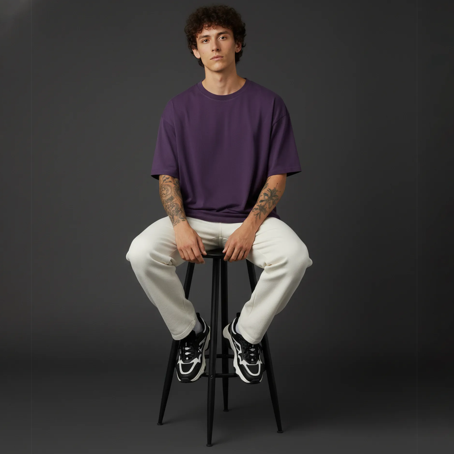 Person sitting on a black stool wearing a purple shirt and white pants against a dark background
