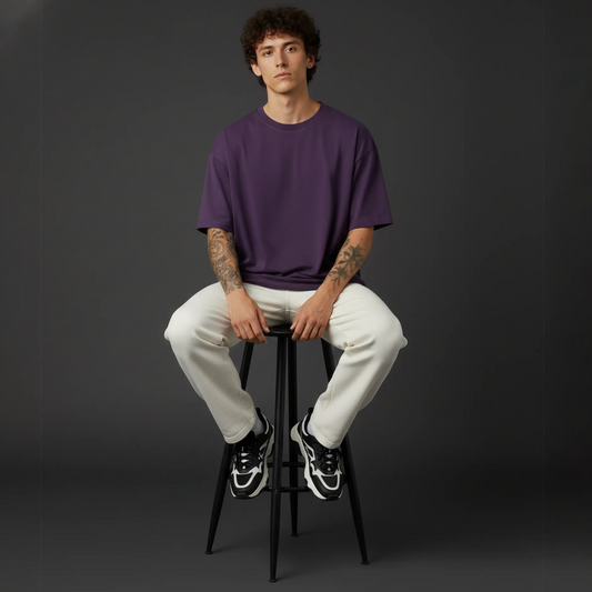 Person sitting on a black stool wearing a purple shirt and white pants against a dark background