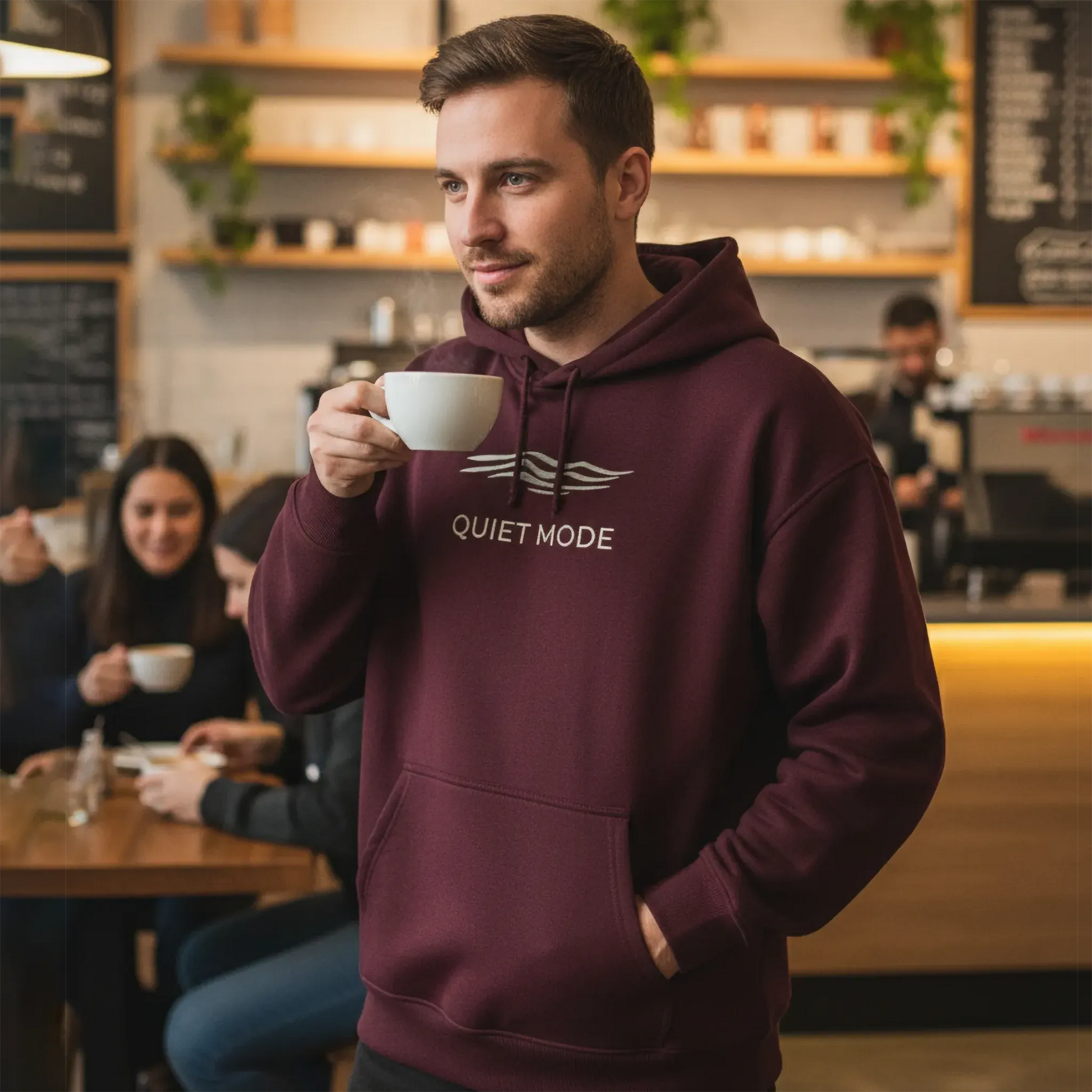 Man in a maroon hoodie with 'Quiet Mode' text holding a cup in a cafe.